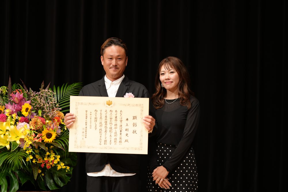 建設ジュニアマスターの表彰状を持って奥様と記念撮影をする井上大工